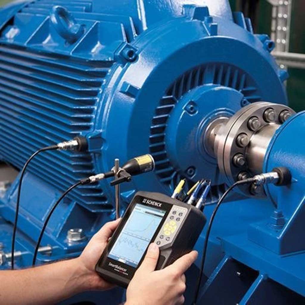 Hands holding a portable diagnostic device connected with cables to a large blue industrial motor for vibration analysis.