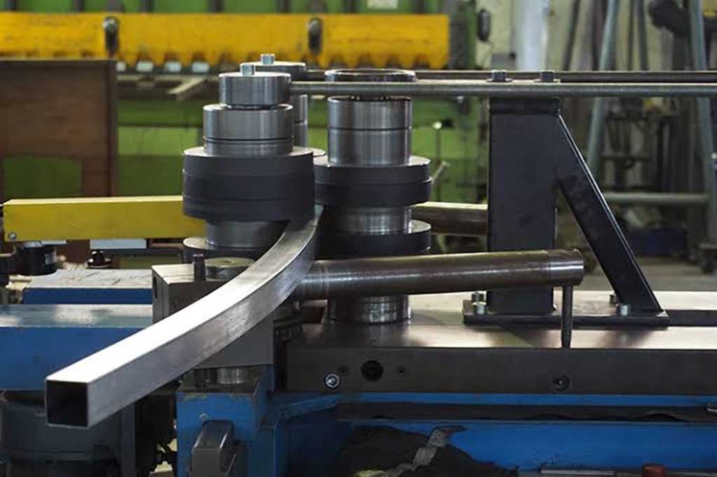 Industrial machine bending a rectangular metal tube inside a workshop.