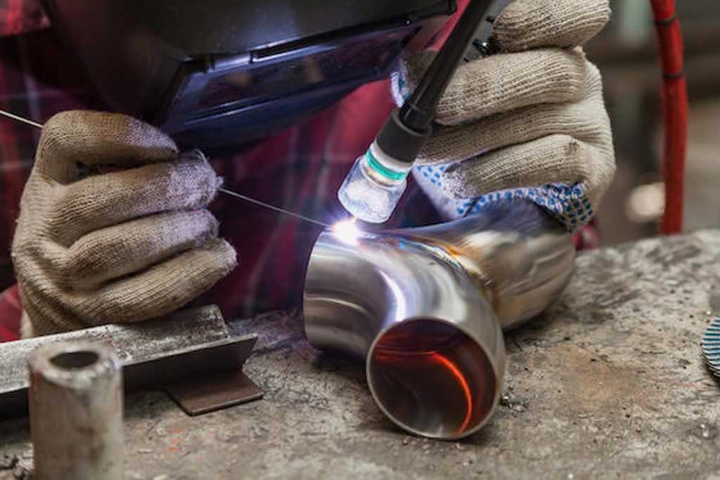 Close-up of a person welding a stainless steel pipe joint while wearing protective gloves and a welding helmet.