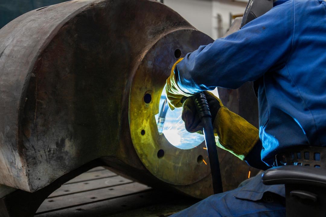 Worker in protective gloves and welding mask welding inside a large metal part with sparks flying.