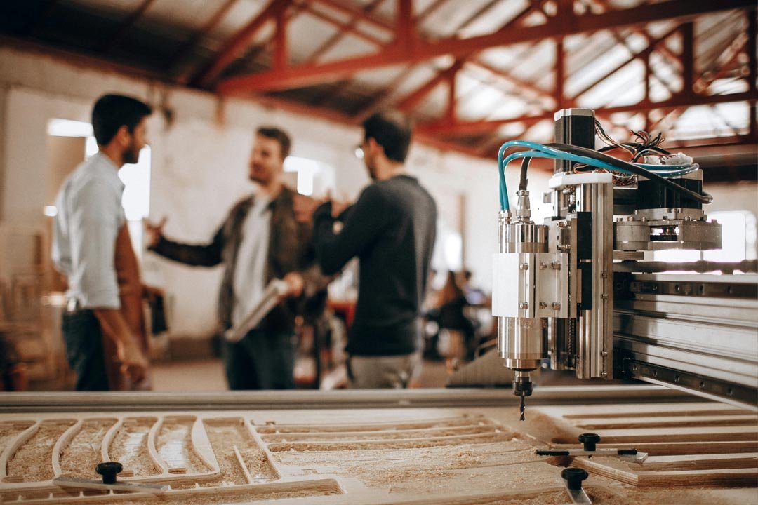Close-up of a CNC machine carving wood with three blurred men discussing in the background inside a workshop.