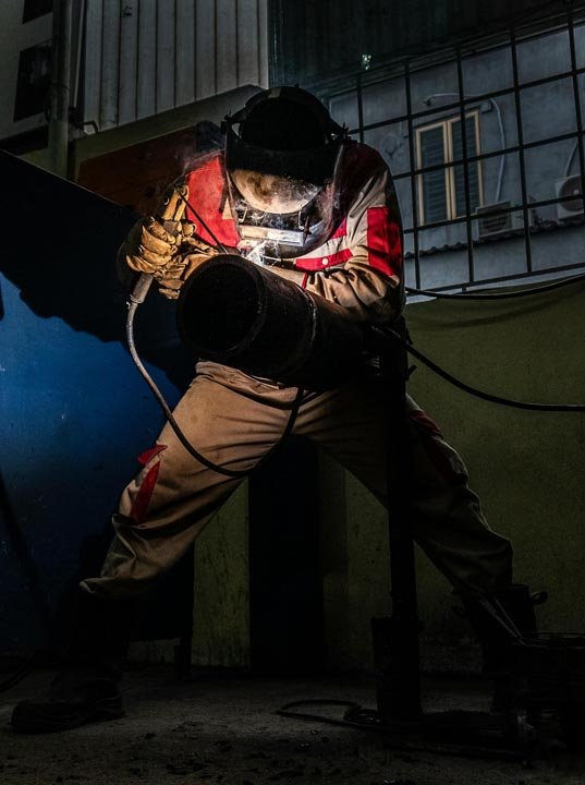 Person wearing protective gear welding a large pipe in a dark industrial setting.