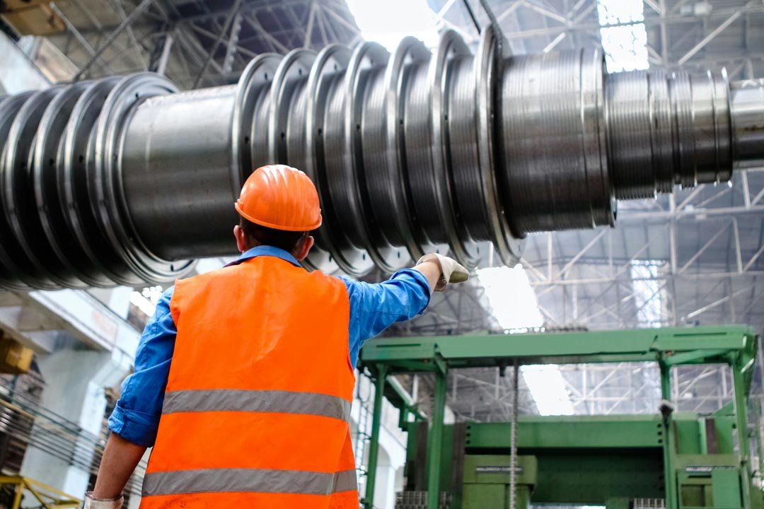 Worker in orange safety vest and helmet pointing at large industrial metal machinery inside a factory.