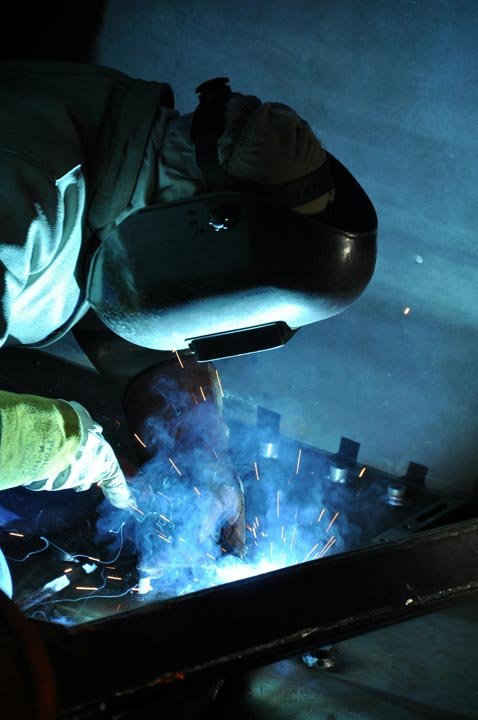Person wearing protective welding helmet and gloves welding metal with bright sparks and smoke.