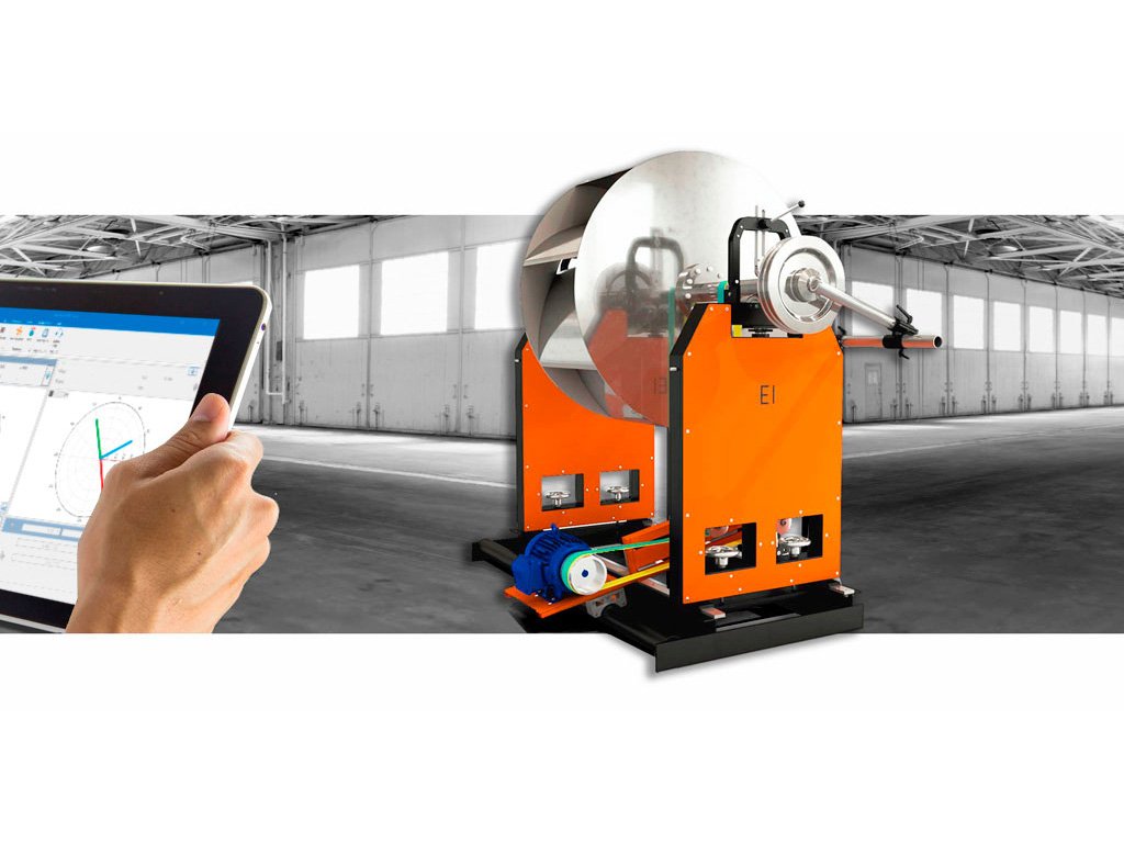 Hand holding a tablet displaying technical data next to an orange industrial machine with a large metal coil in a spacious warehouse.