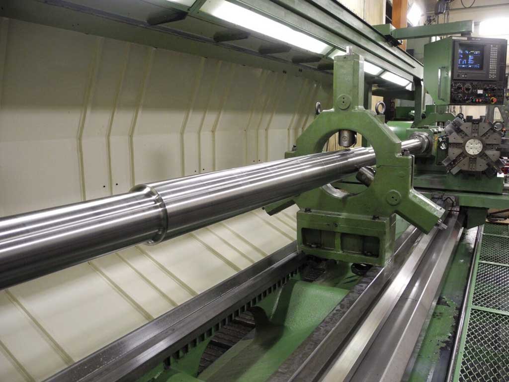 Industrial CNC lathe machine holding a long, polished metal cylindrical workpiece inside a factory setting.